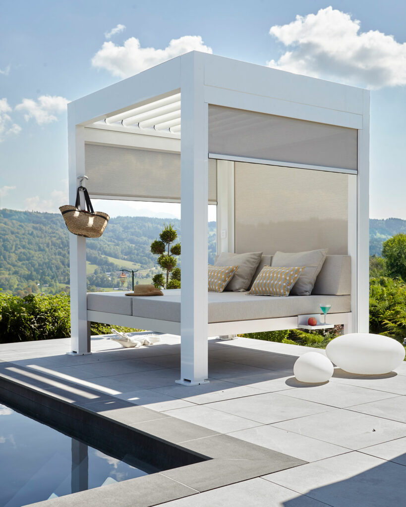 Lit extérieur 2 places intégré dans une structure de pergola bioclimatique équipée de stores latéraux occultants et installée au bord d'une piscine.