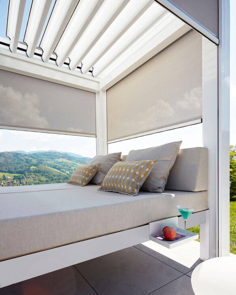 Vue rapprochée sur le lit outdoor bioclimatique qui comporte un matelas et des oreillers dont le tissus est très qualitatif et décoratif. Plateau métallique de côté en accessoire sur le quel sont posé un verre de jus de fruit et une pomme. Un chapeau de paille est accroché sur un patère installé sur un des montants verticaux de la structure