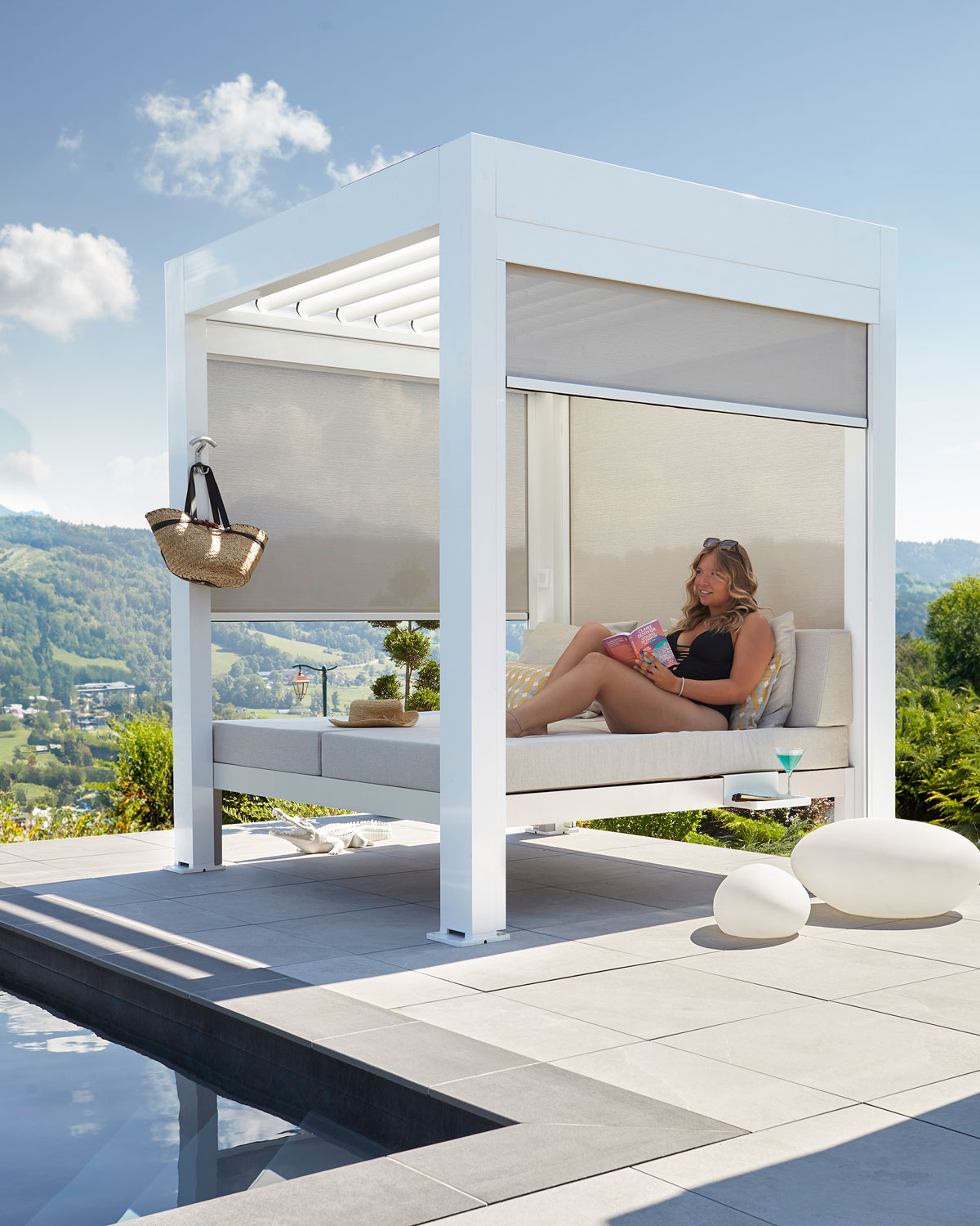 Lit extérieur 2 places intégré dans une structure de pergola bioclimatique équipée de stores latéraux occultants et installée au bord d'une piscine. Une jeune femme y est installée et profite de la convivialité que procure le lit outdoor